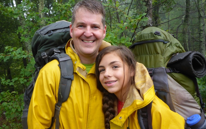 A parent and child pose for a photo in a green wooded area. Both are wearing rain jackets and backpacks.